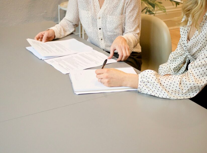 woman signing on white printer paper beside woman about to touch the documents