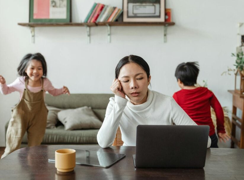 A tired mother working on a laptop while her children play around indoors, highlighting remote work challenges.